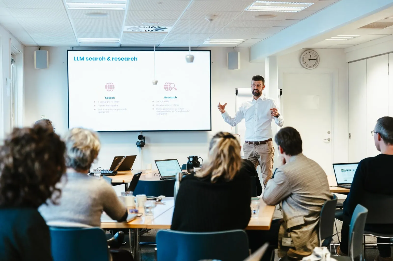 A trainer presents at a large screen to a class of participants sitting with laptops.